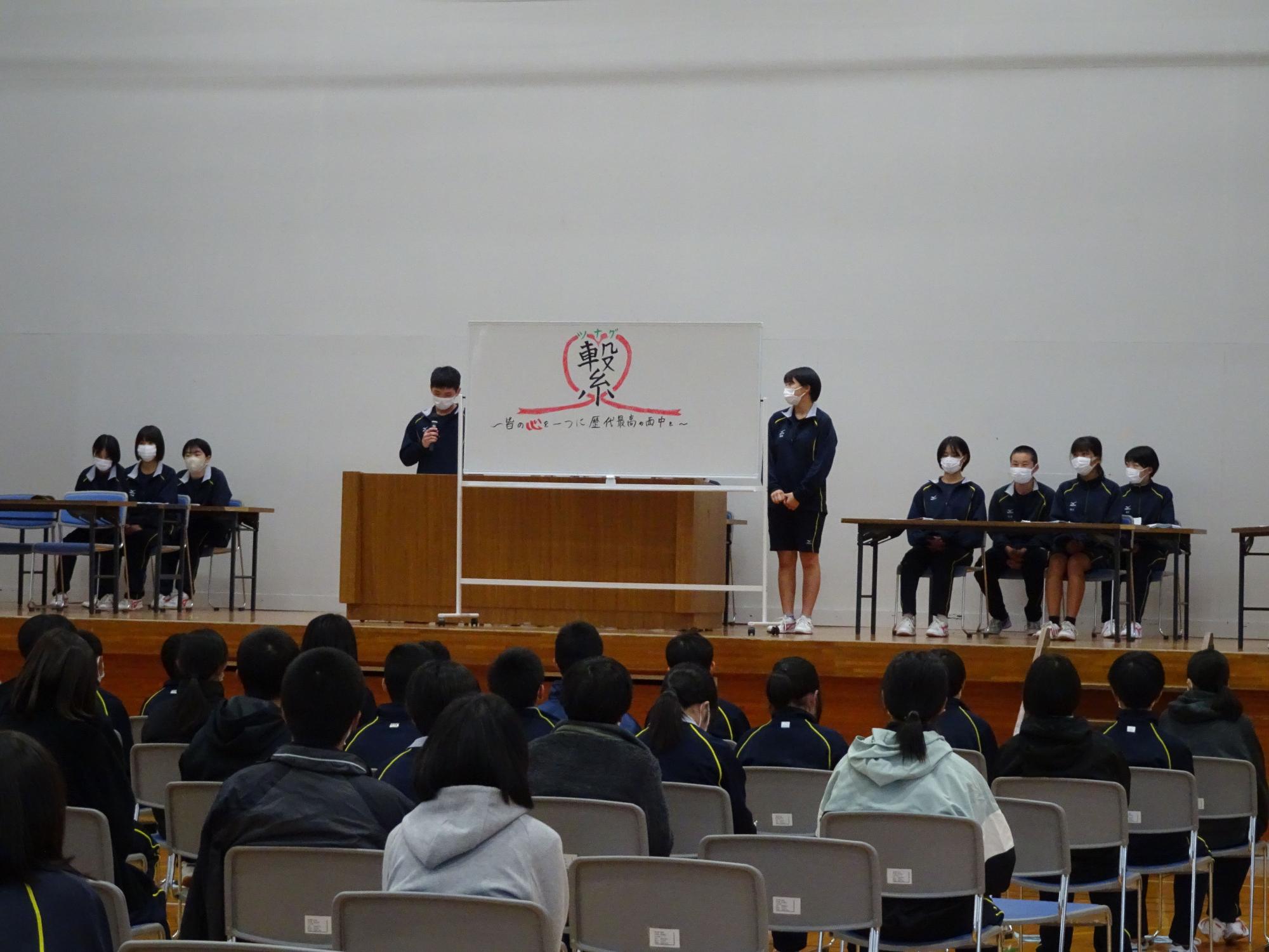 繋ぐと書かれたホワイトボードの横に立って話している生徒の写真