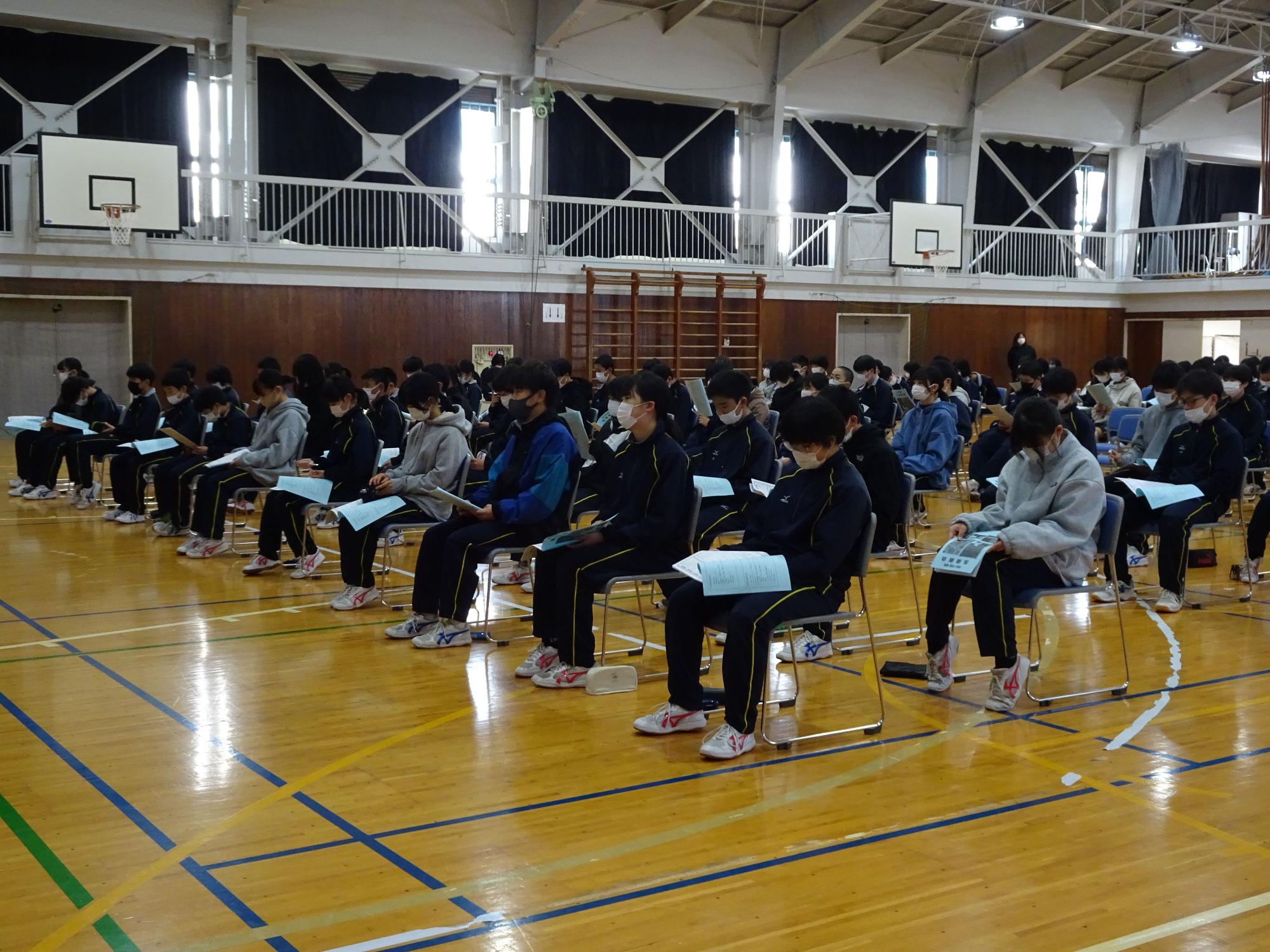 体育館で椅子に座り手元の資料を読んでいる生徒たちの写真