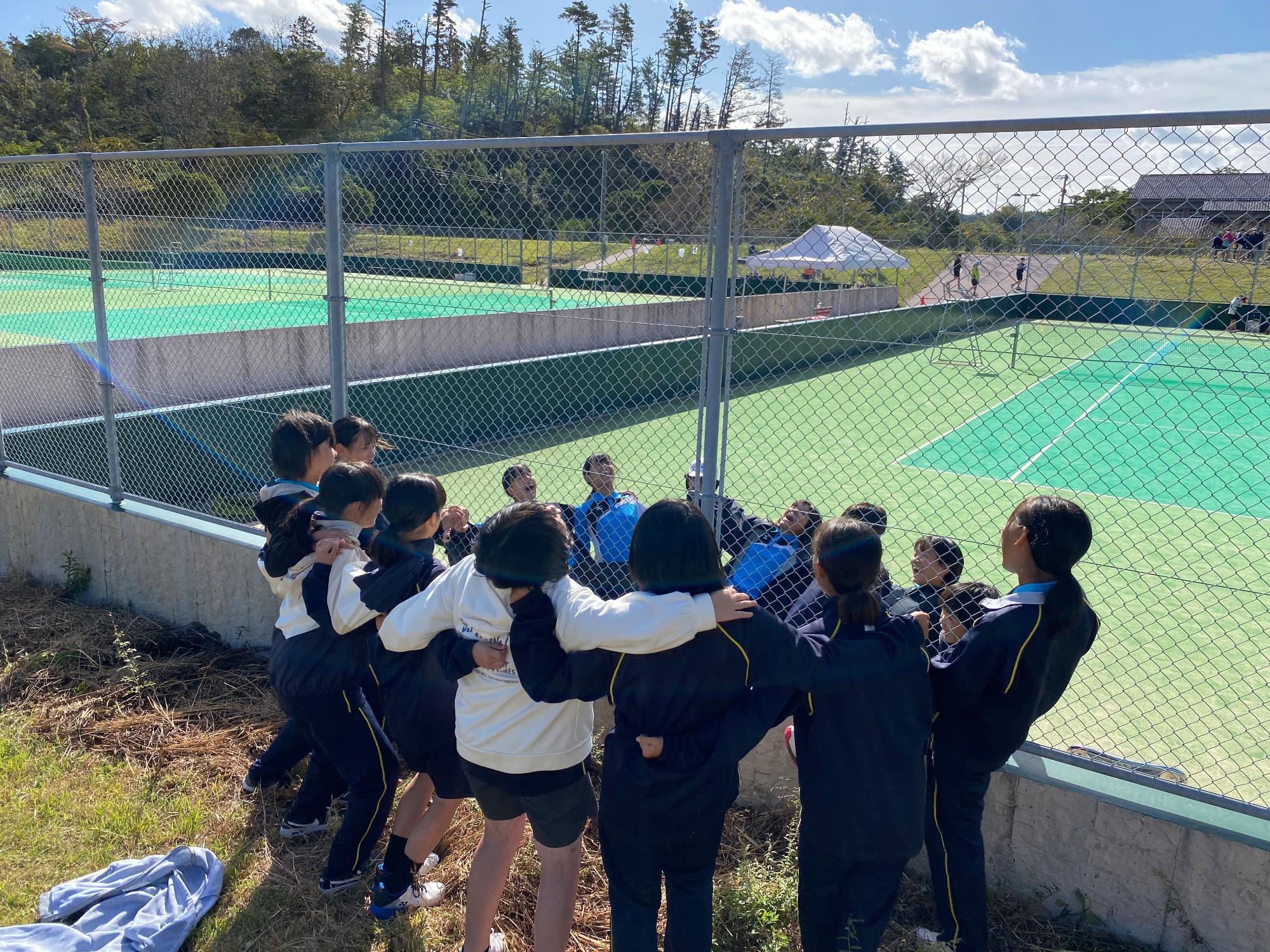 テニスコートのフェンスをはさんで円陣を組む生徒たちの写真