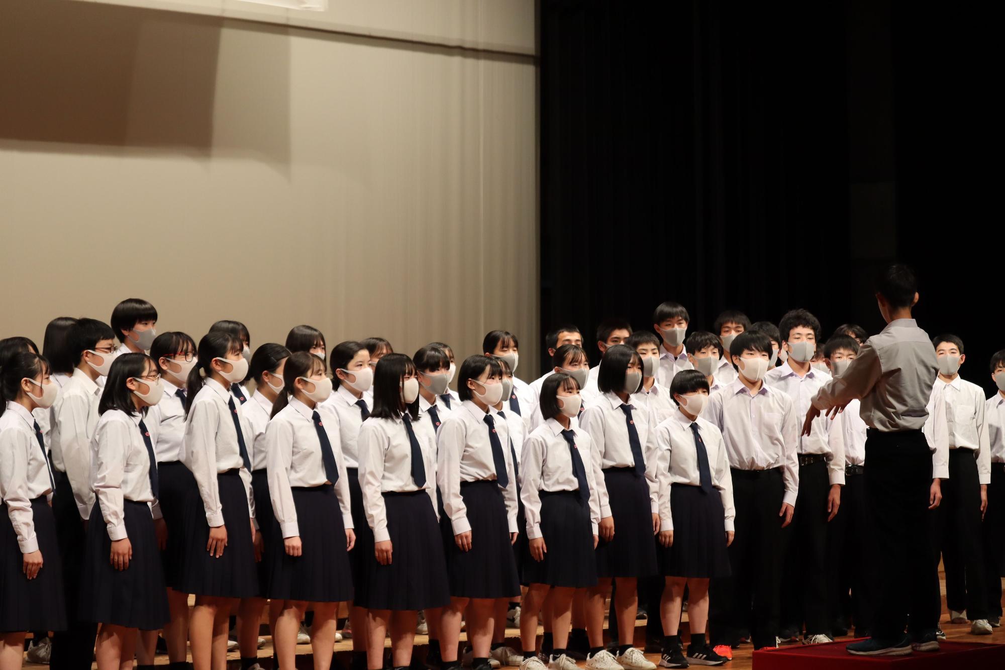 舞台上で男性の指揮者に合わせて合唱する生徒たちの写真