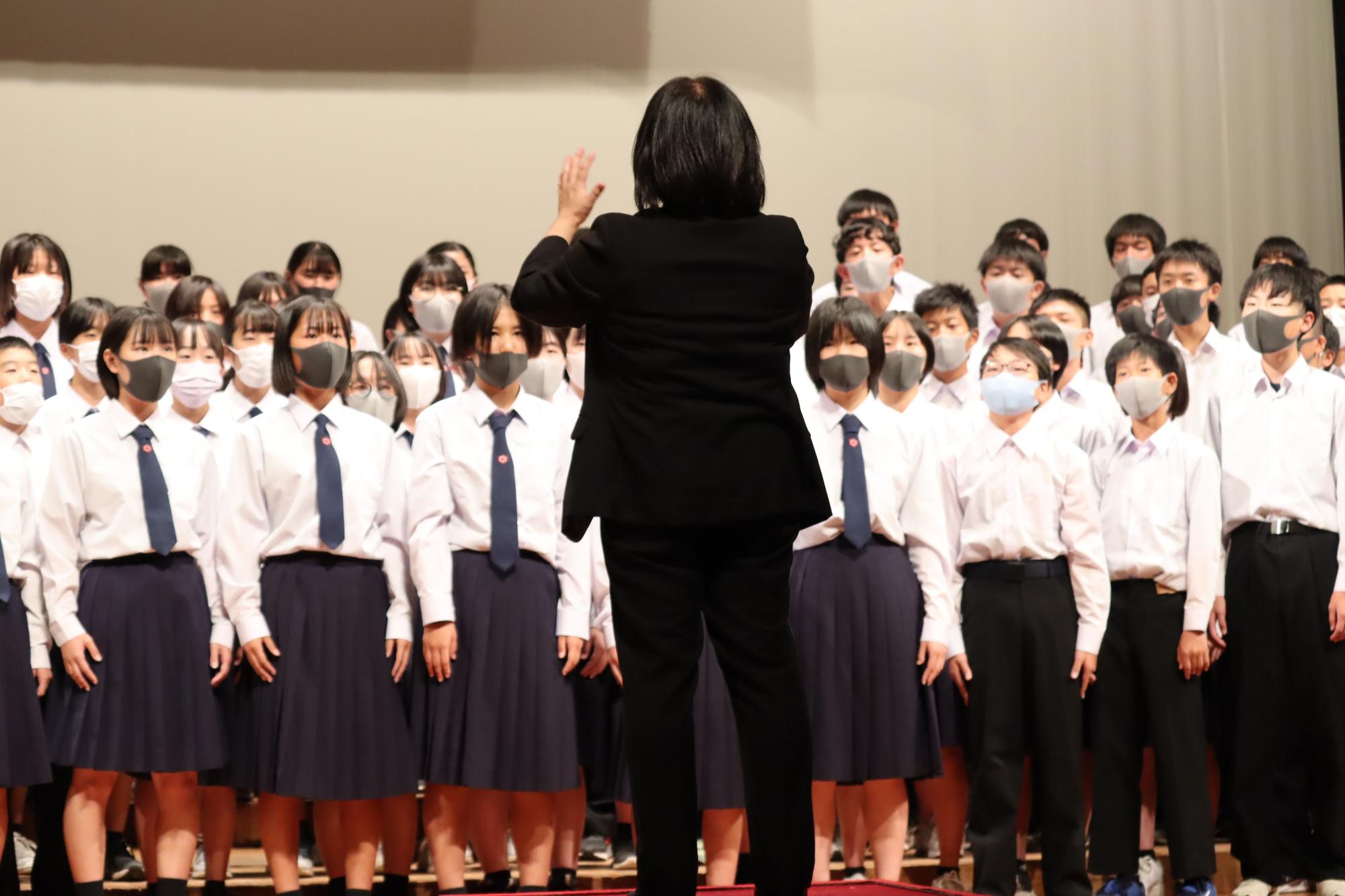 舞台上で女性の指揮者に合わせて合唱する生徒たちの写真