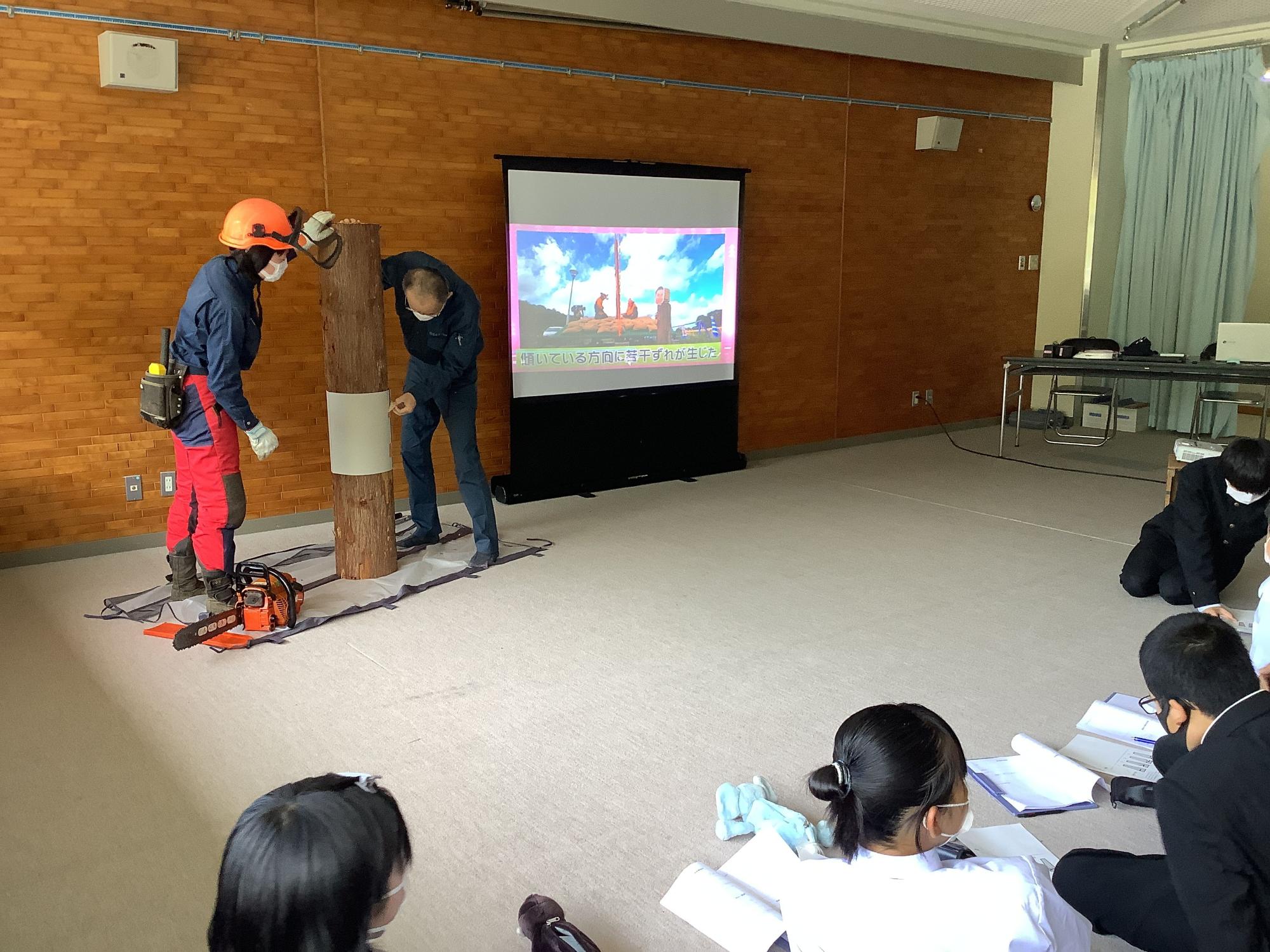 モニター横に丸太を置き実演する2人の男性と座ってみている生徒たちの写真