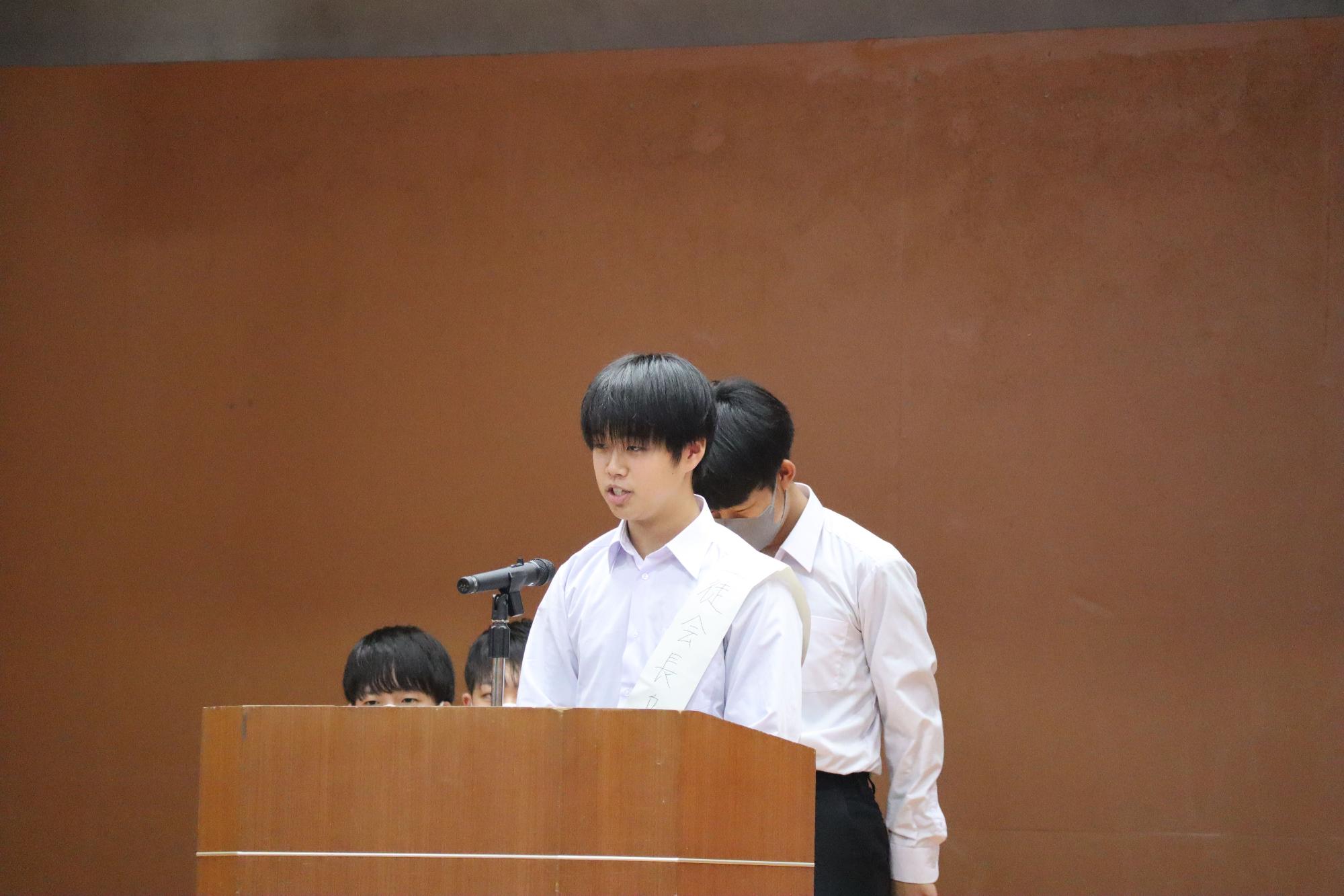マイクの置かれた演壇の前に立って話している男子生徒の写真