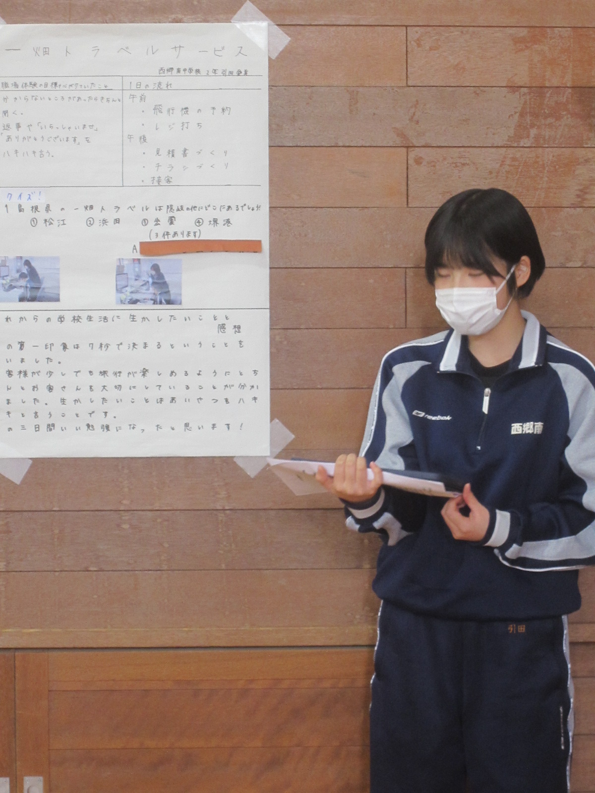 壁に貼られたポスターの横に立ち発表している女子生徒の写真