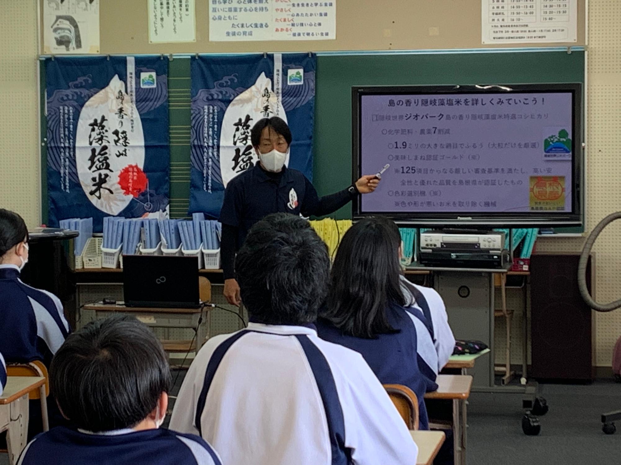 黒板の前に置かれたモニターを指して教鞭を執る先生と話しを聞いている生徒の写真写真