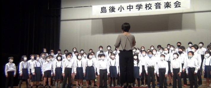 体育館の舞台で先生が指揮をして児童たちが合唱をしている写真