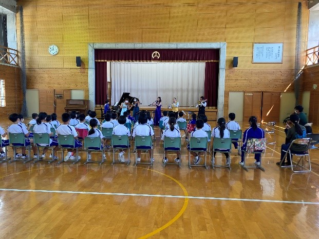 体育館の中央で児童たちが椅子に座って演奏を鑑賞している写真