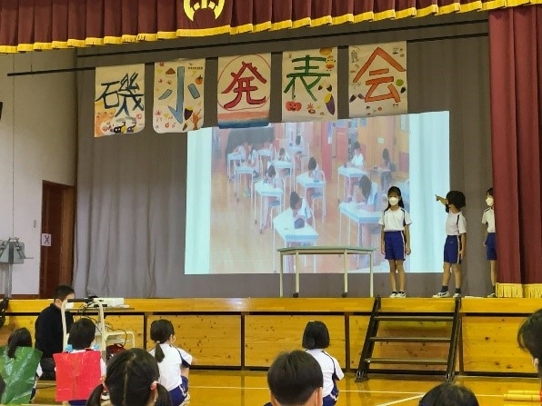 体育館の舞台で児童たちが映像を使って発表している写真