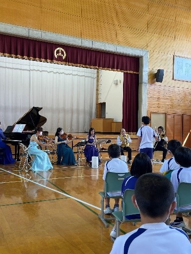 体育館の舞台前で児童が楽器の演奏を聴いている写真