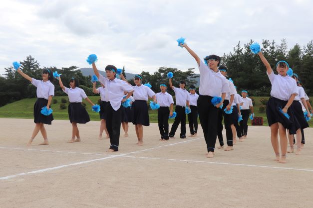 運動場で生徒たちが青いポンポンを持ってダンスをしている写真