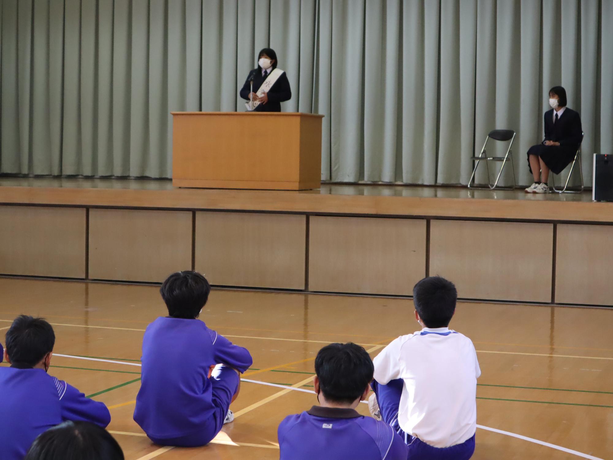体育館でたすきをかけた生徒が壇上で話をしている写真
