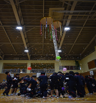 体育館で児童たちの頭上に紙吹雪が舞い、くす玉が割れる瞬間を写した写真