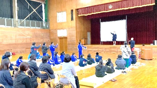 体育館で児童たちがステージ上の制服姿の生徒の発表を座って見ている写真