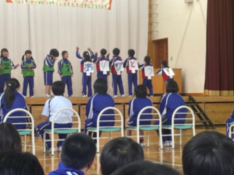 体育館のステージで児童たちが「ありがとう」と書いた文字を背に発表している写真