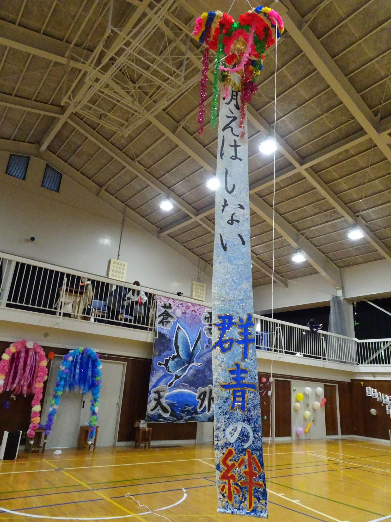 体育館の天井から「消えはしない群青の絆」と文字が書かれた大きなくす玉が垂れ下がっている写真