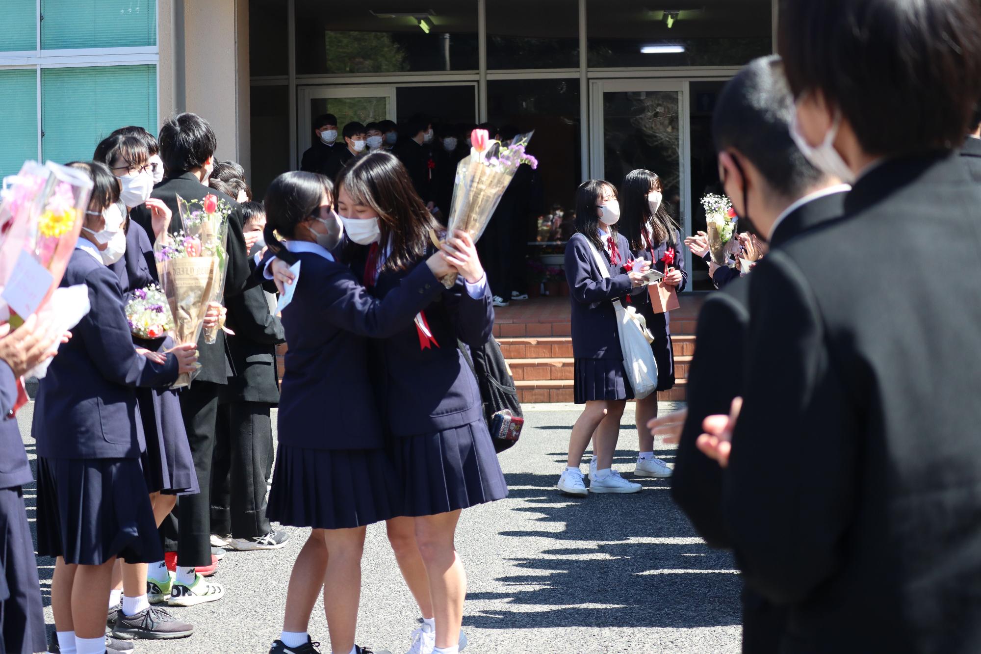 校舎の前で卒業生たちが花束を持って抱き合っている写真