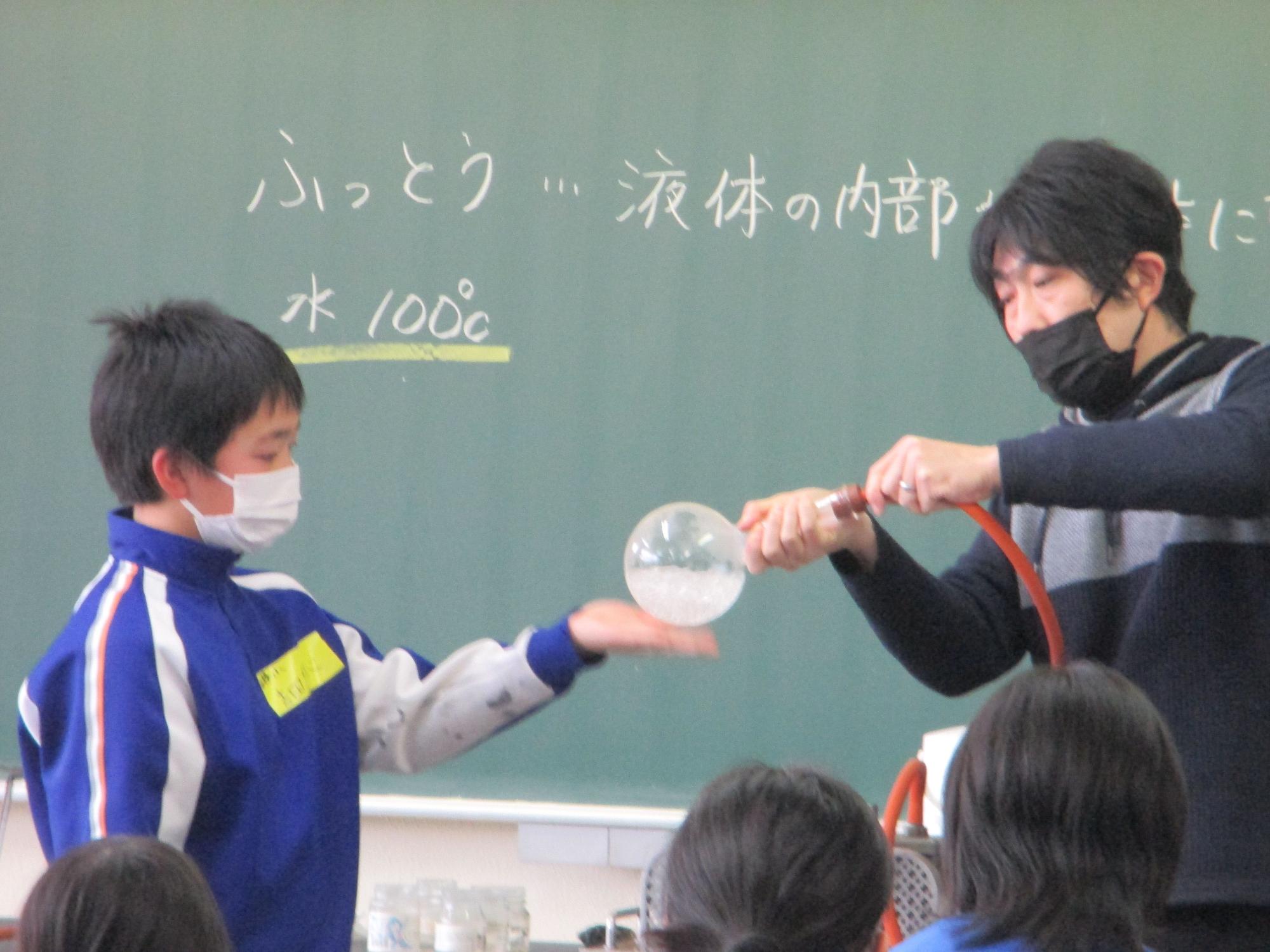 理科室で先生と生徒が黒板の前で実験をしている写真