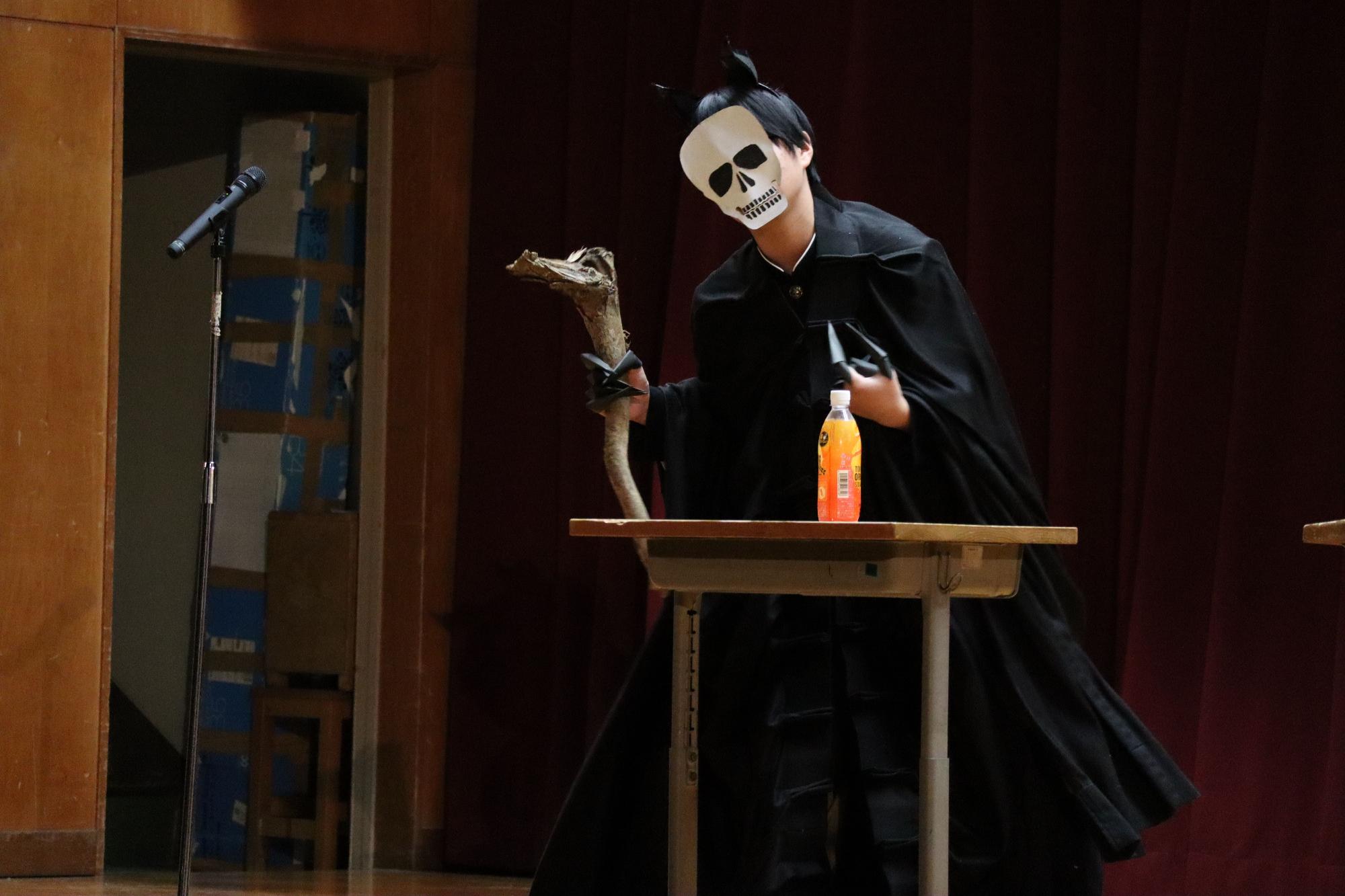 体育館の舞台で仮面をつけた生徒が劇の発表をしている写真