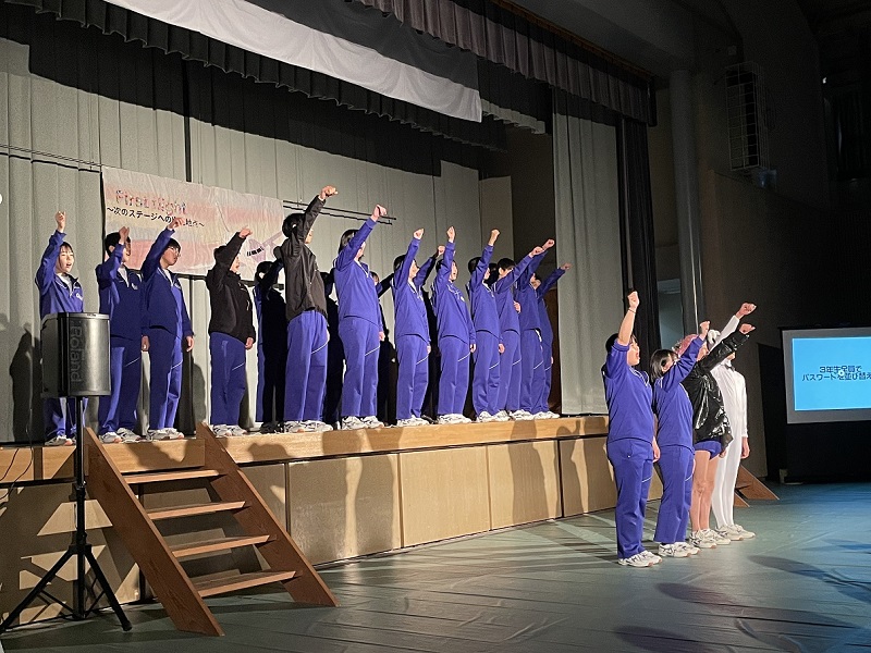 体育館の舞台で生徒たちがこぶしを挙げて発表をしている写真