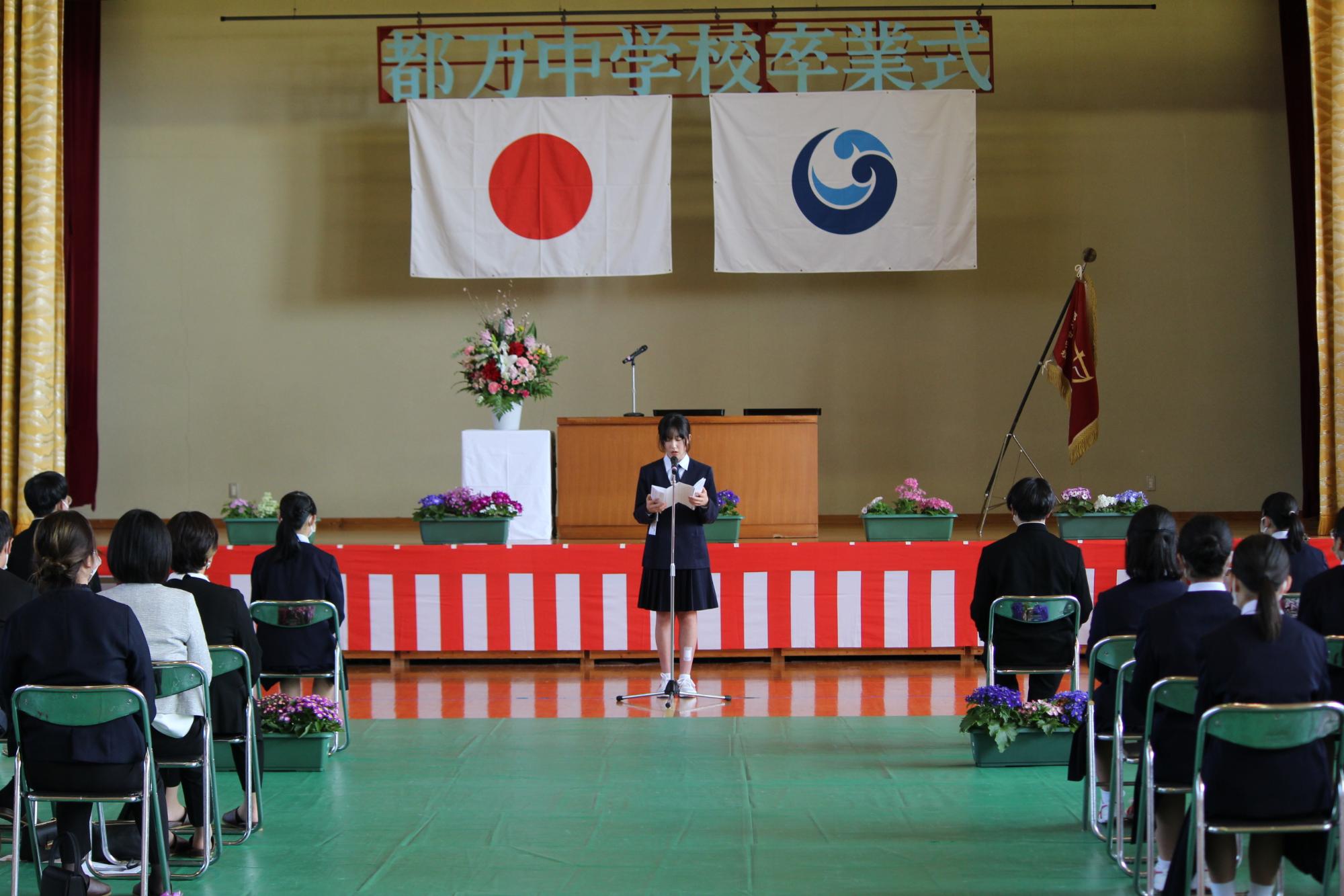 体育館で行われている卒業式で代表生徒が前に立って話している写真