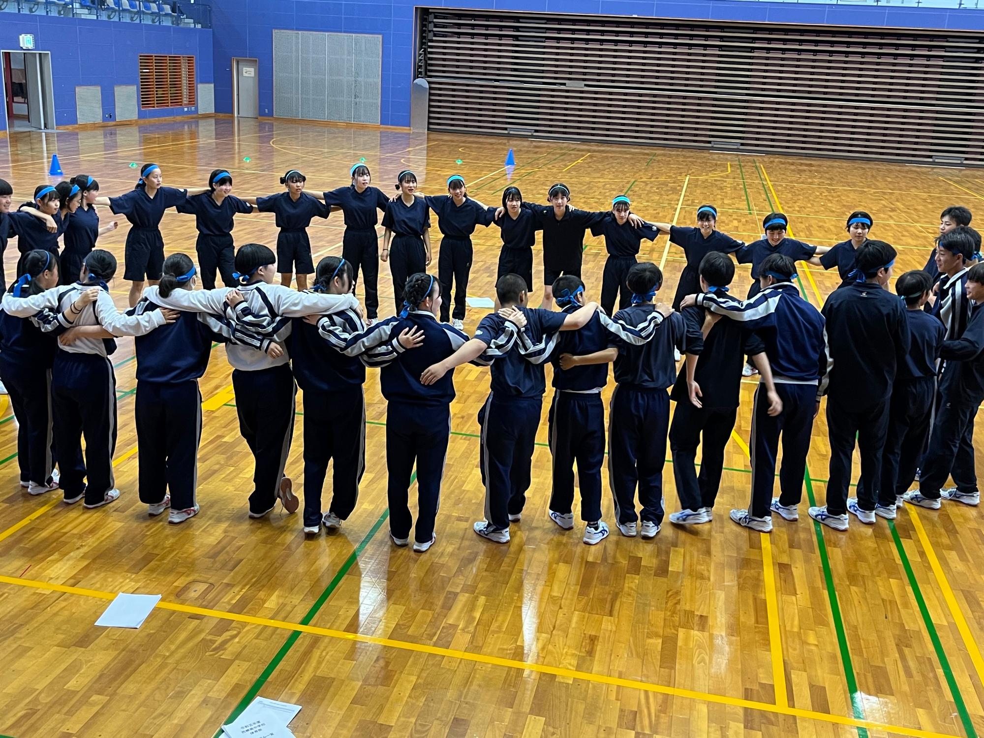 体育館で生徒たちが円になって肩を組み団結している写真
