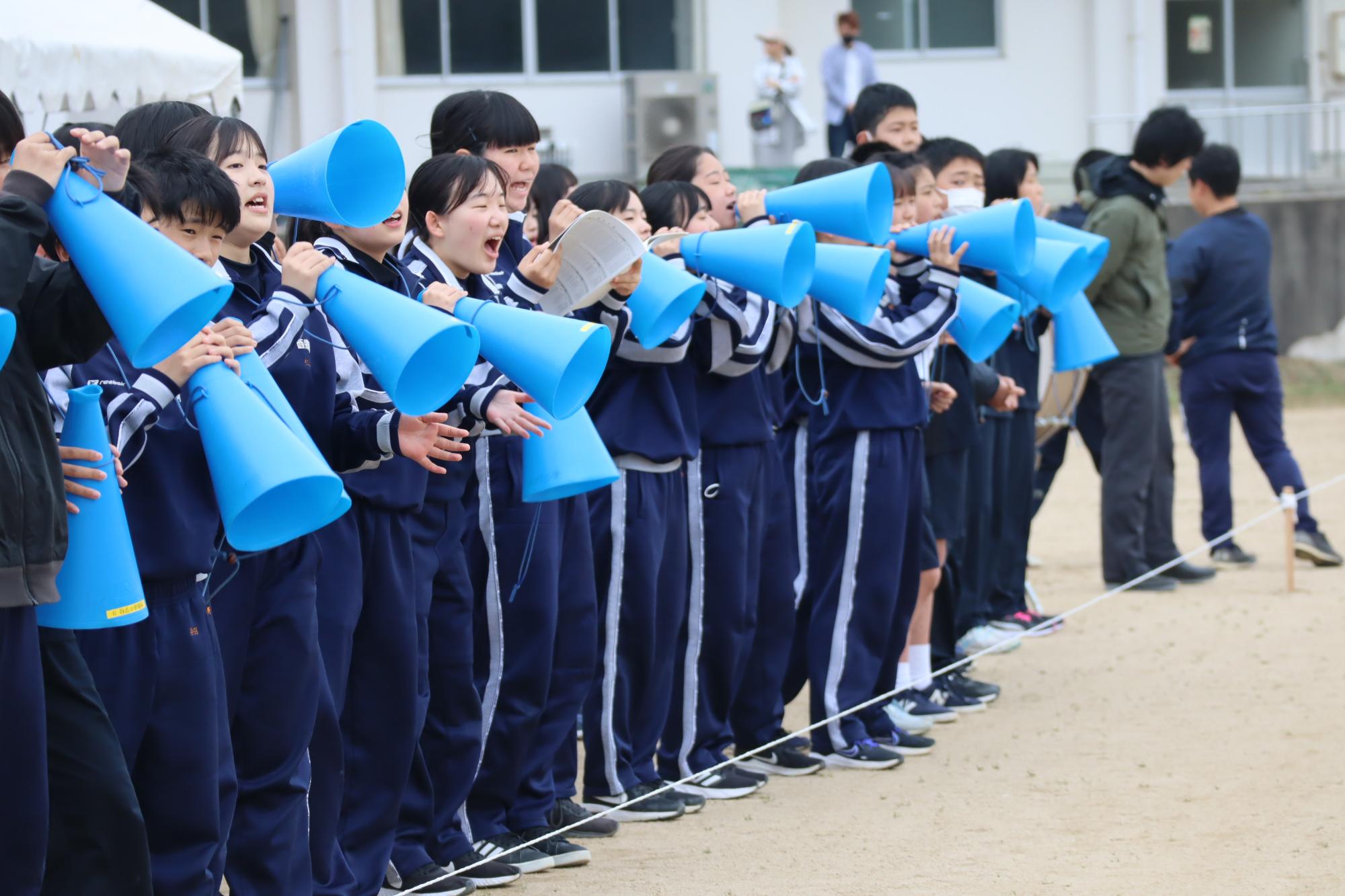 グラウンドで生徒たちが青いメガホンを使って応援している写真