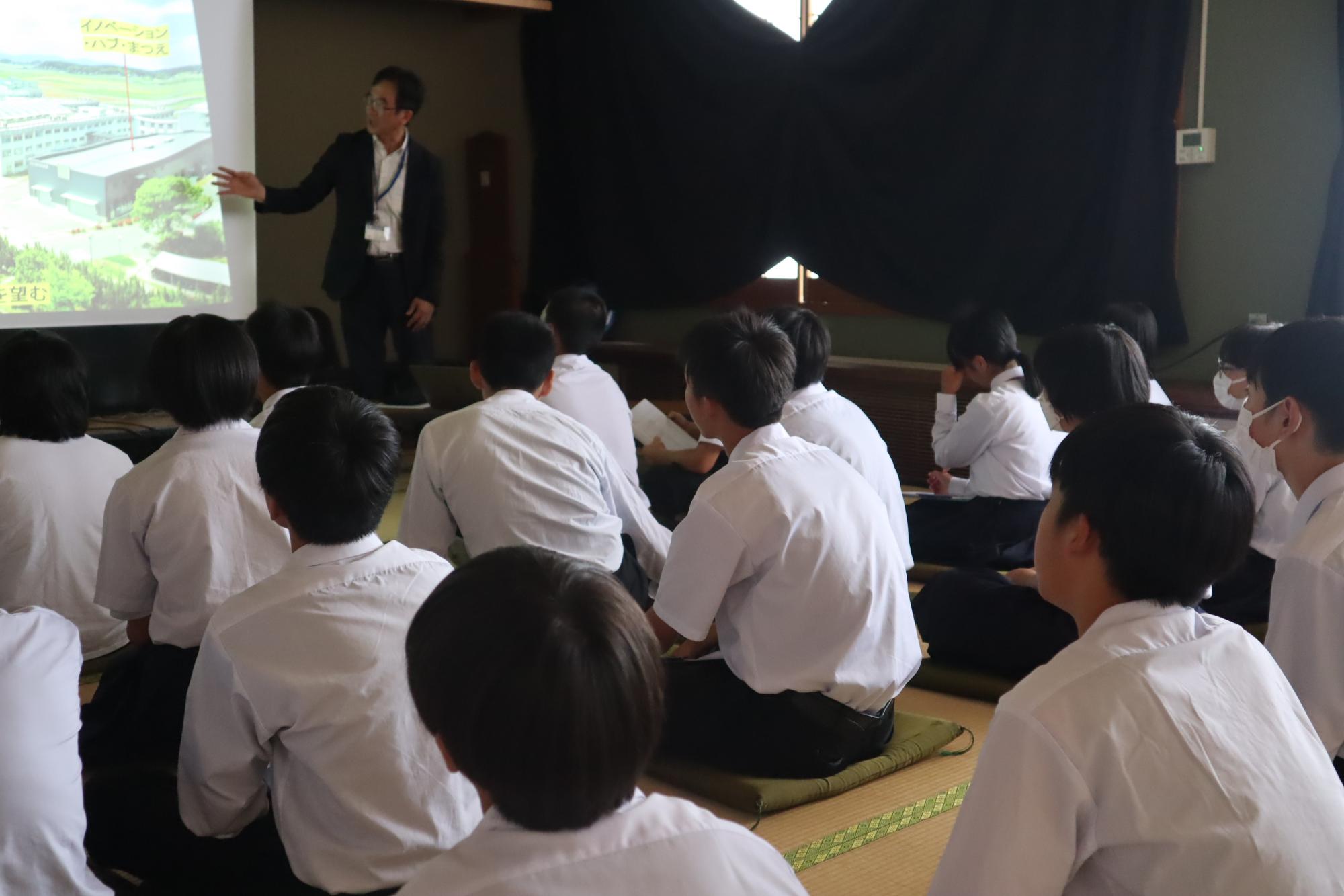 講師が前方でスクリーンを使いながらプレゼンを行い、生徒たちが真剣に聞いている写真