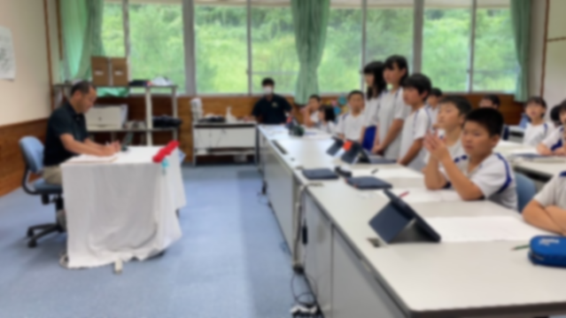 教室で児童たちが白い机の前に立ち先生の話を聞いている写真