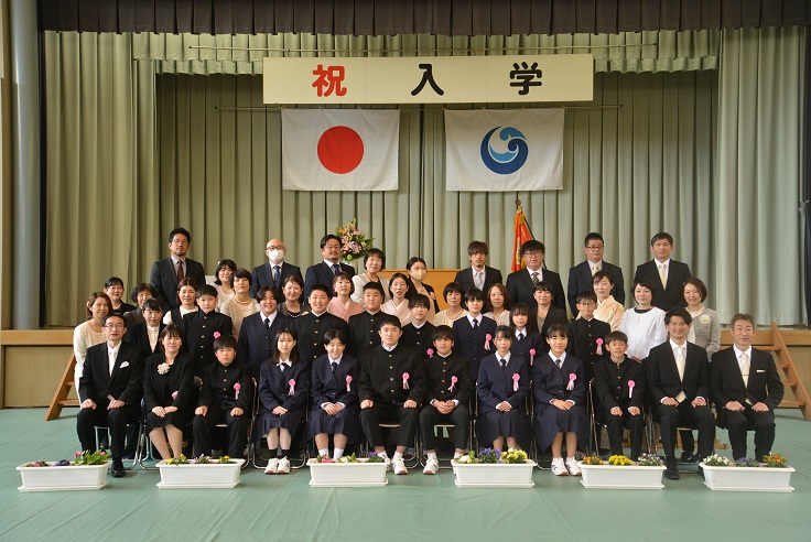 体育館の舞台で新入生と教職員、保護者がそろって入学式の記念撮影をしている写真