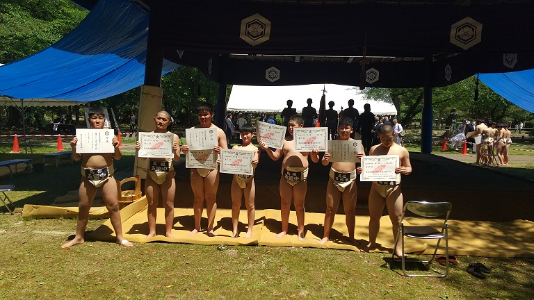 屋外の土俵の前で相撲大会に出場した生徒たちが賞状を持って並んでいる写真