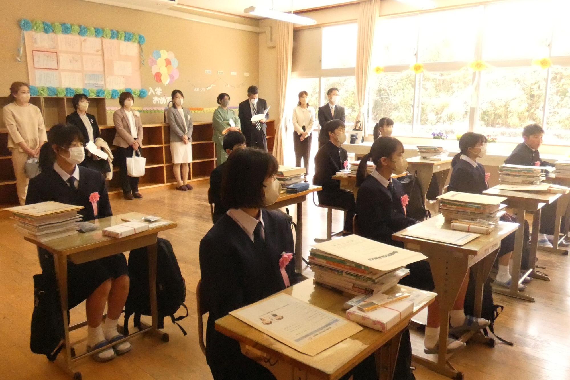 教室で新入生が席に座り、保護者が後ろに立って先生の話を聞いている写真