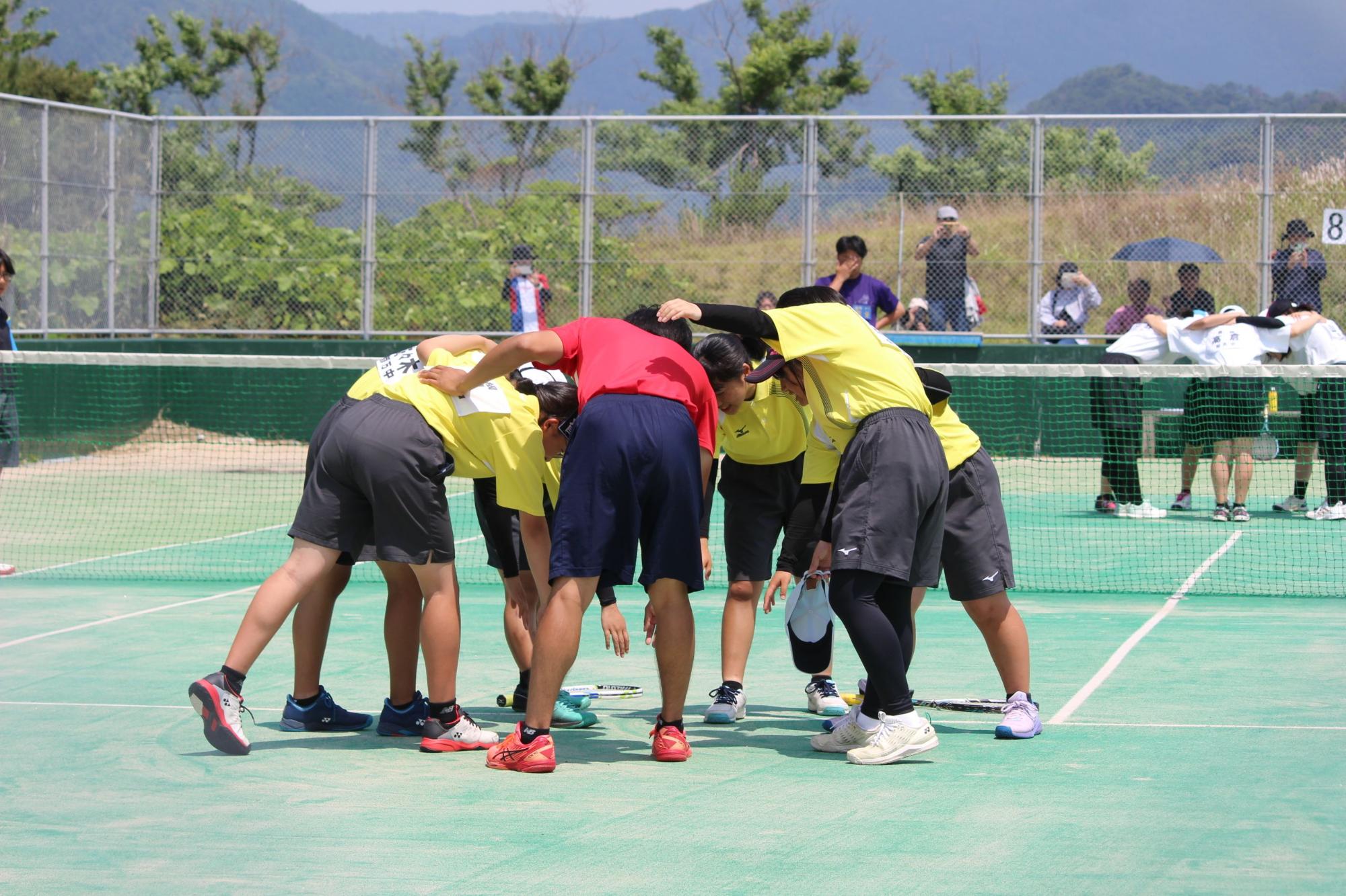 テニスコートで黄色のユニフォームの生徒たちが円陣を組んでいる写真