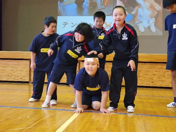体育館の檀上前で4人の男子児童が決めポーズをしている写真