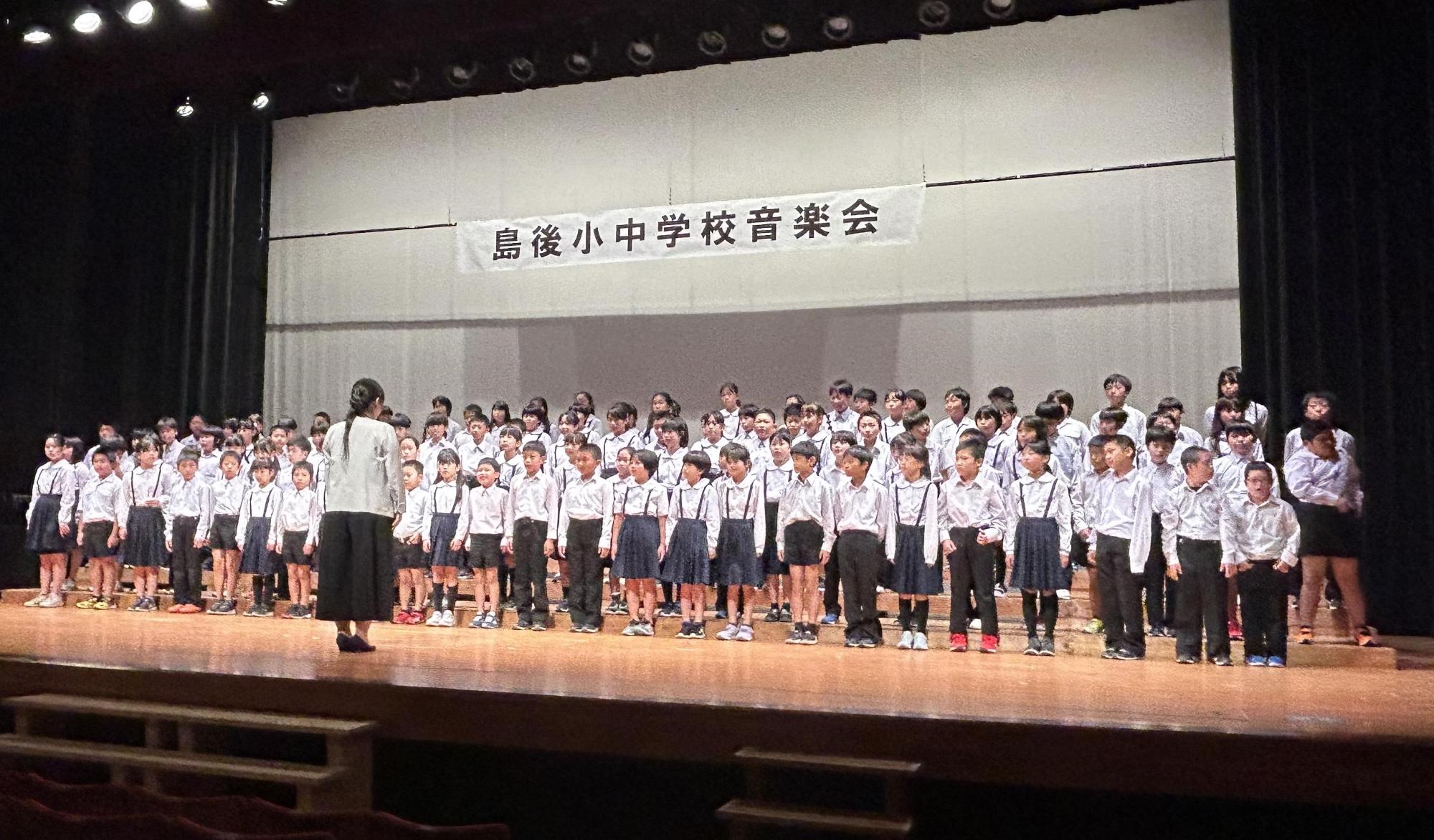 舞台上で小学生と中学生が4列に並んで合唱をしている写真
