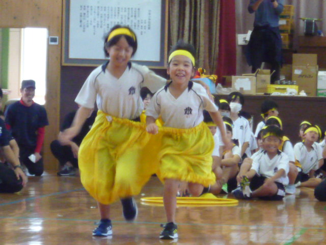 黄色のハチマキを着用した二人の児童が体育館で並んで走っている様子の写真
