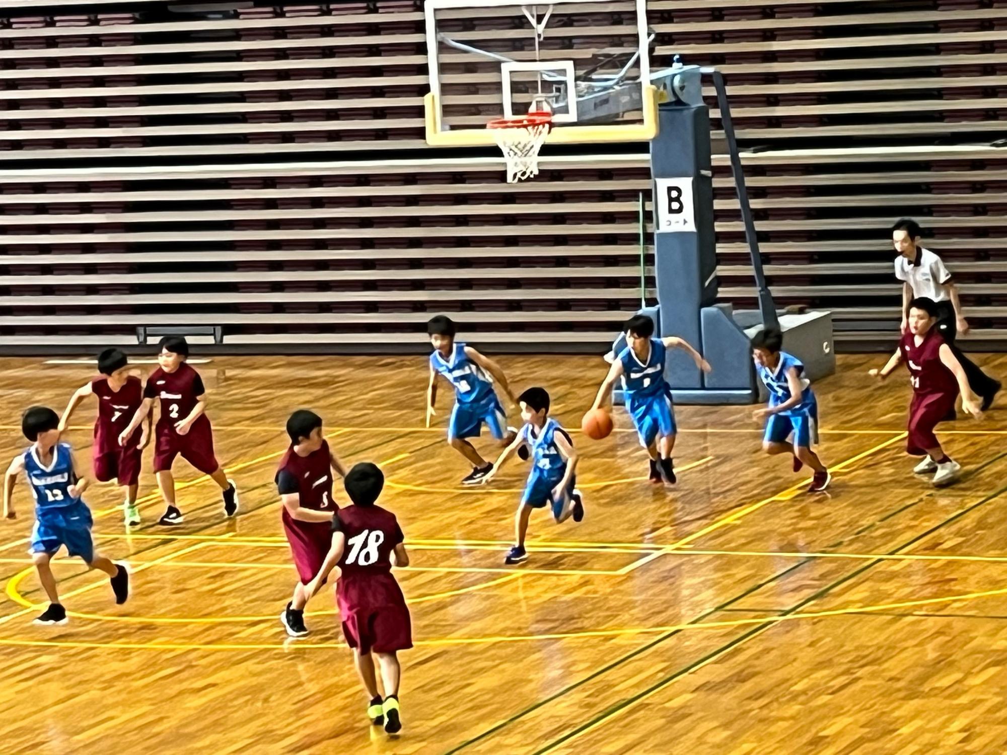 複数の児童が赤色のユニフォームを着たチームと青色のユニフォームを着たチームに分かれてバスケットボールの試合をしている写真
