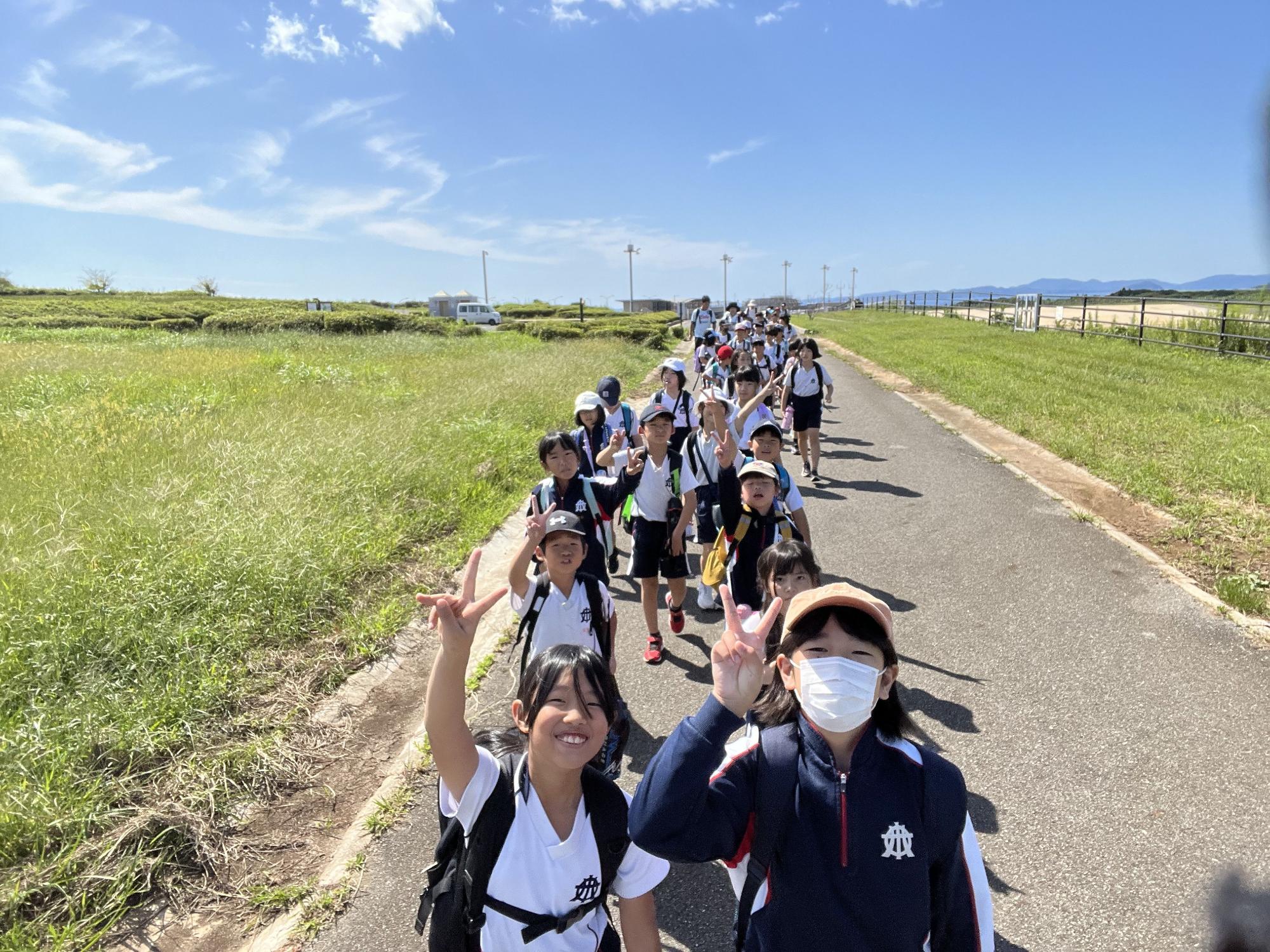 屋外の一本道を一列に並びピースをしている児童を正面から撮影した写真