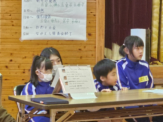 体育館で司会席に座る青いジャージ姿の4人の児童の写真