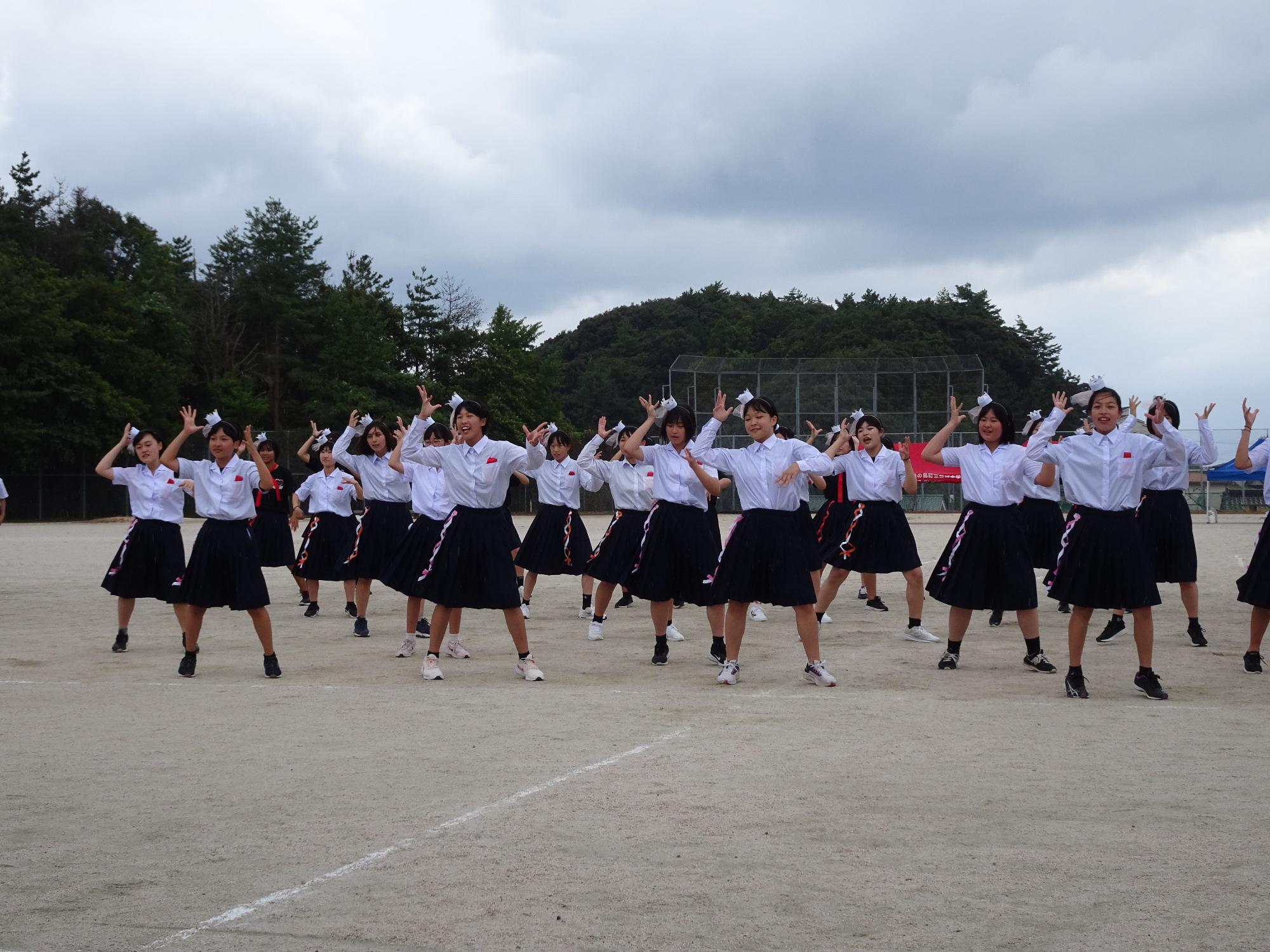 運動場で女子生徒が並んで片手を挙げてダンスを披露する写真