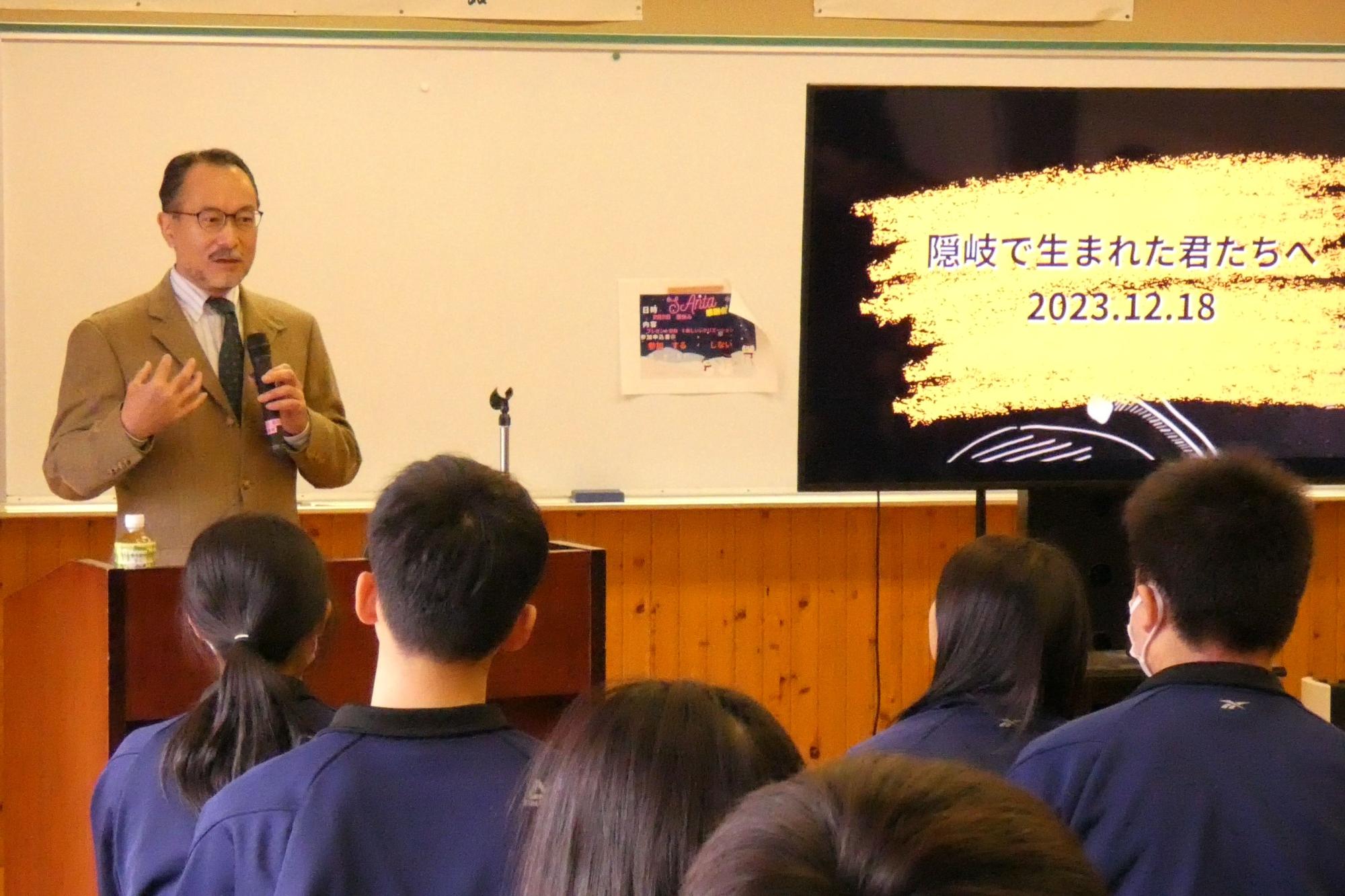 講演室で茶色いスーツを着た講師がマイクを持って生徒に講演をしている写真