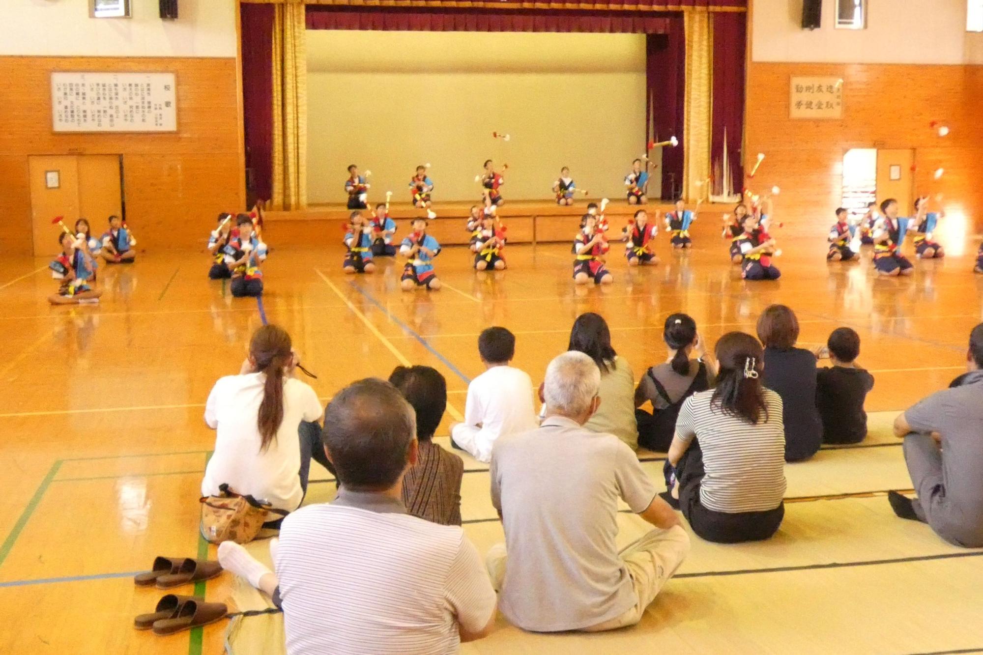 体育館で衣装を着た児童が観客の前で踊りを披露している写真