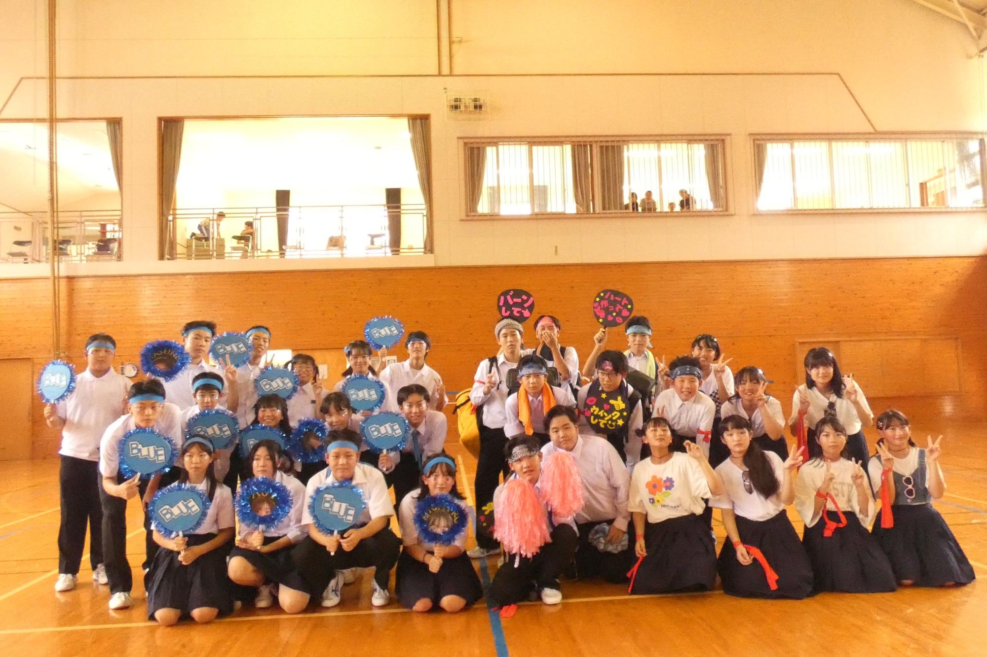 体育館で青いはちまきをつけた生徒たちが団扇を持って集合写真を撮っている写真