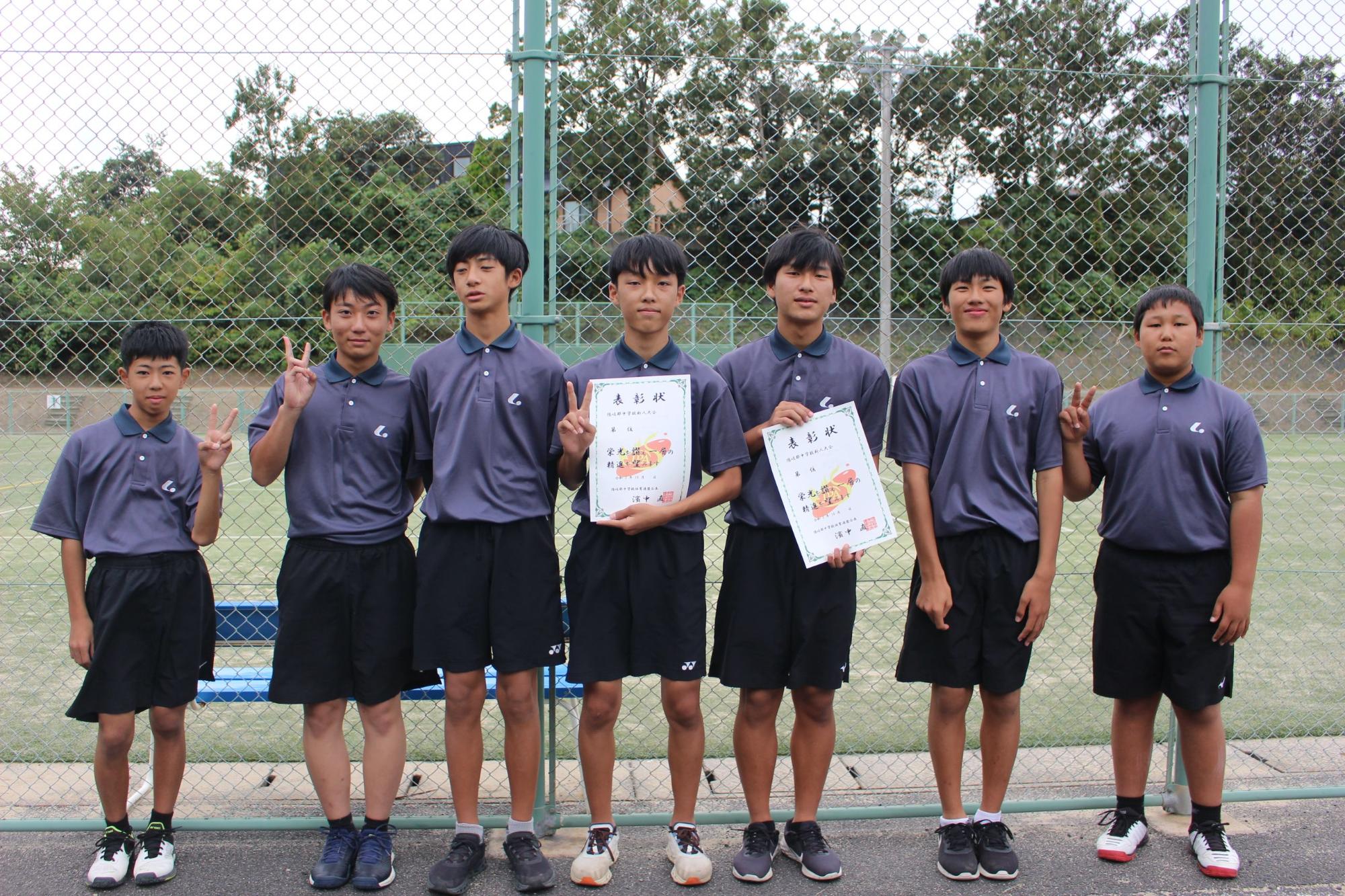 屋外のテニスコート前で男子生徒が表彰状を持って並んでいる写真