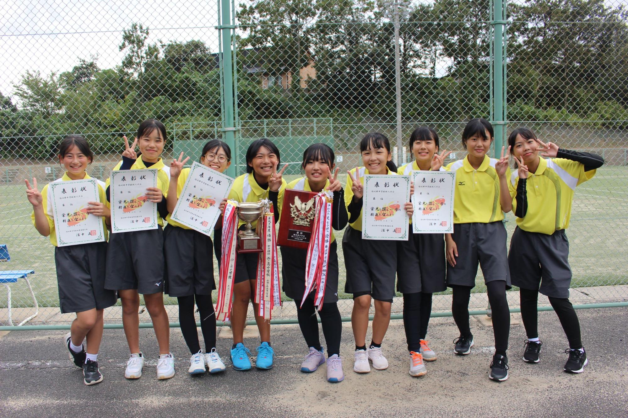 屋外のテニスコート前で女子生徒が表彰状とトロフィーを持って並んでいる写真
