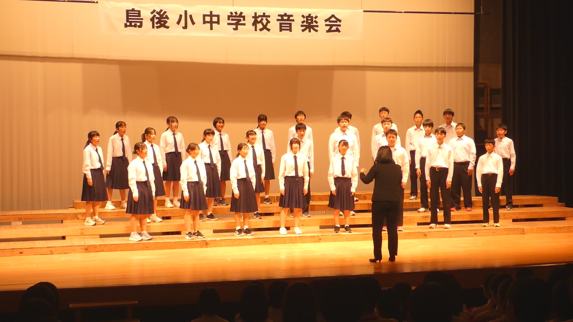 ステージ上で制服姿の生徒たちが指揮者の方向を向き合唱をしている写真