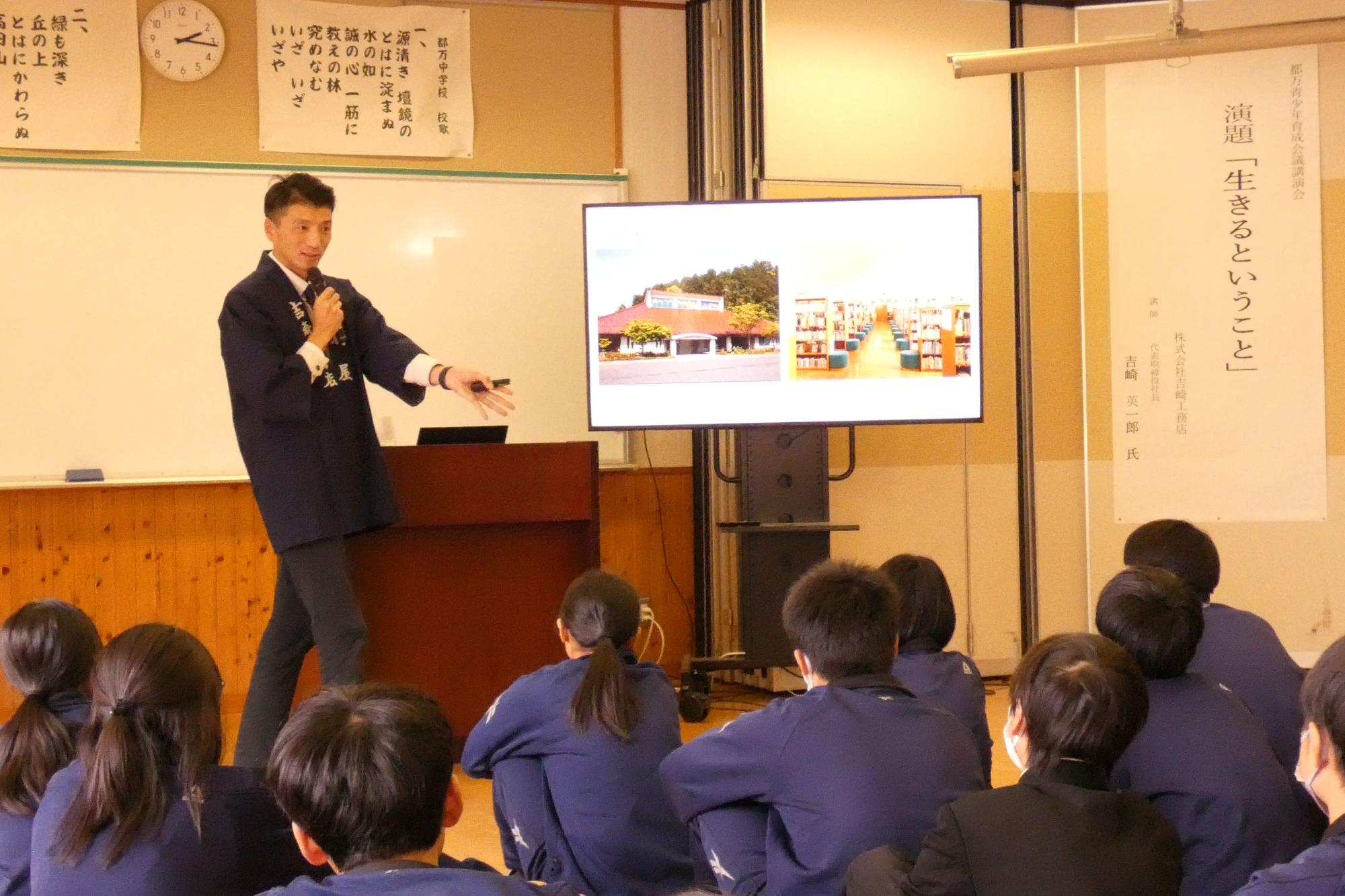 講演室で黒法被を着た講師がマイクを持って生徒に講演をしている写真