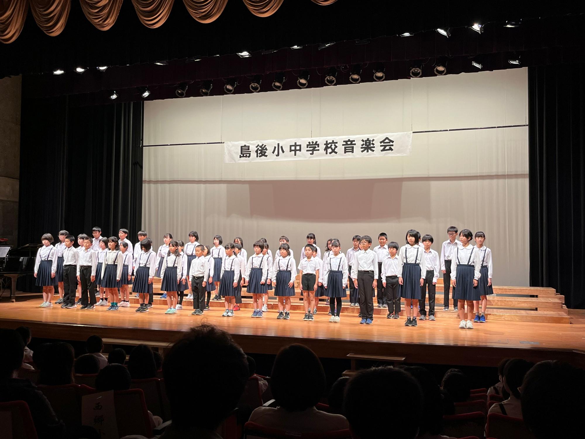 島後小中学校音楽会と書かれた横断幕のある舞台上で整列している数十人の児童たちの写真