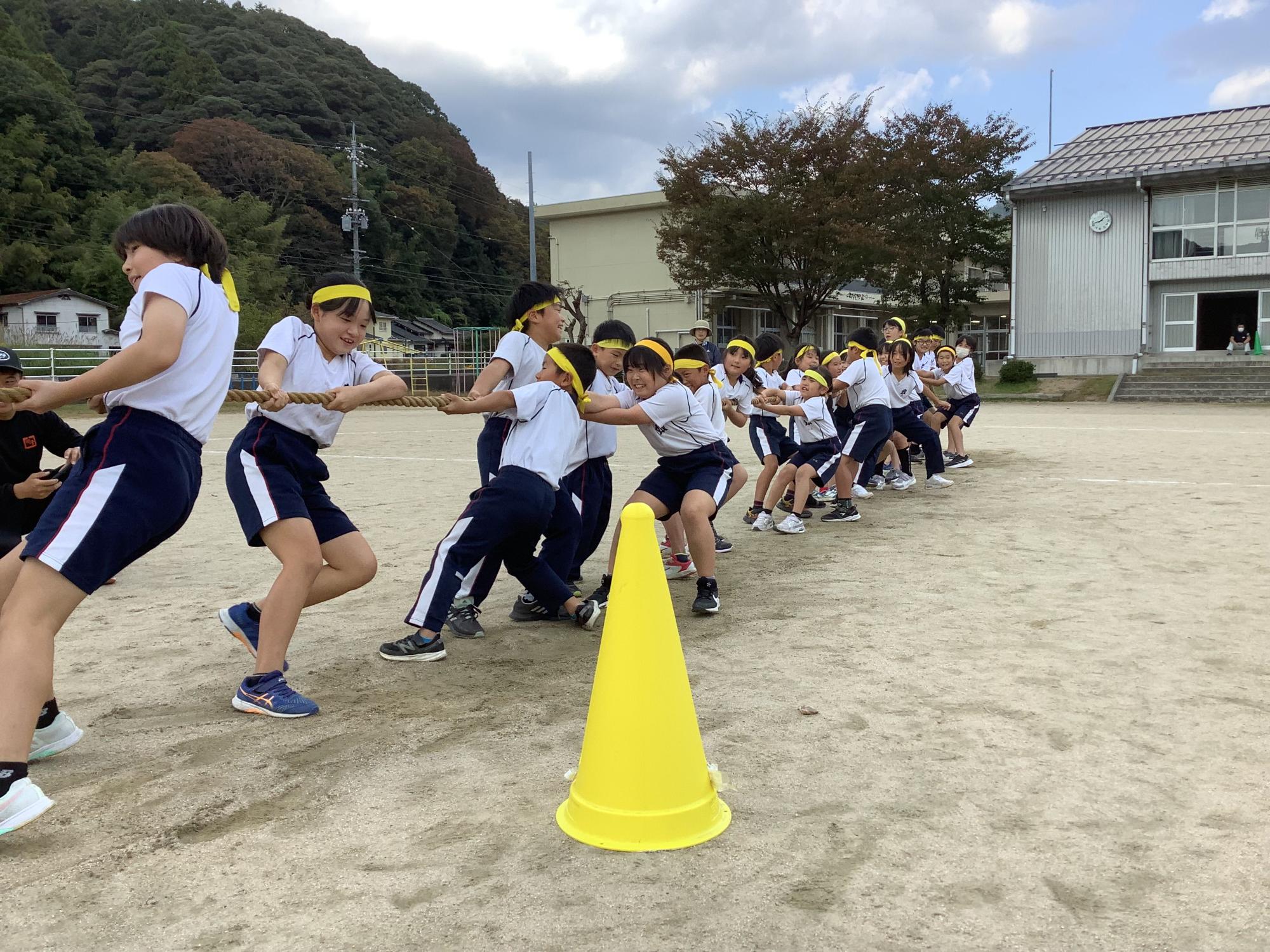 グラウンドで綱引きをする黄色いのハチマキを巻き体操着を着用した複数の児童たちの写真