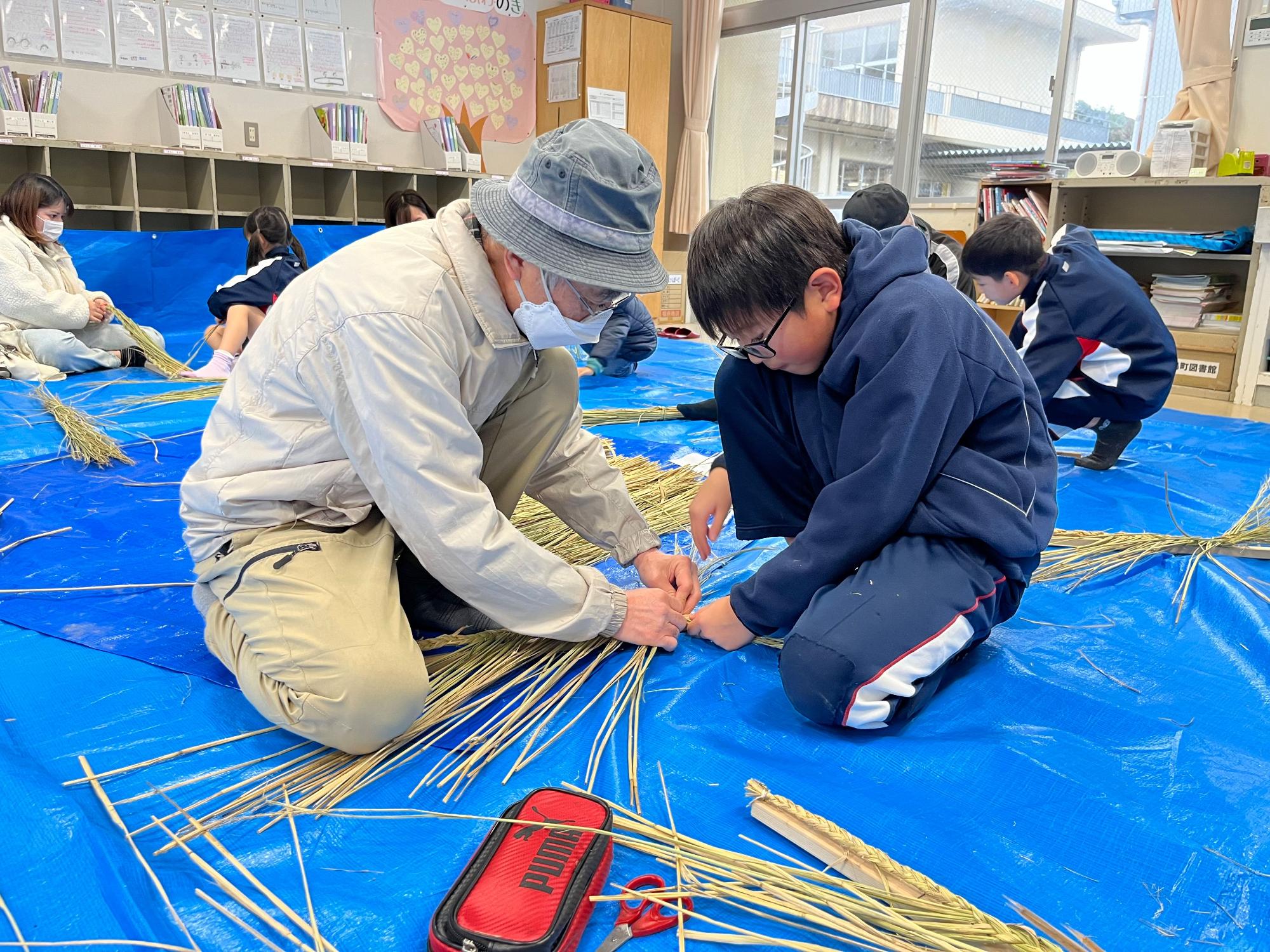 ブルーシートの上で講師と一緒にしめ縄つくりに挑戦している一人の児童の写真