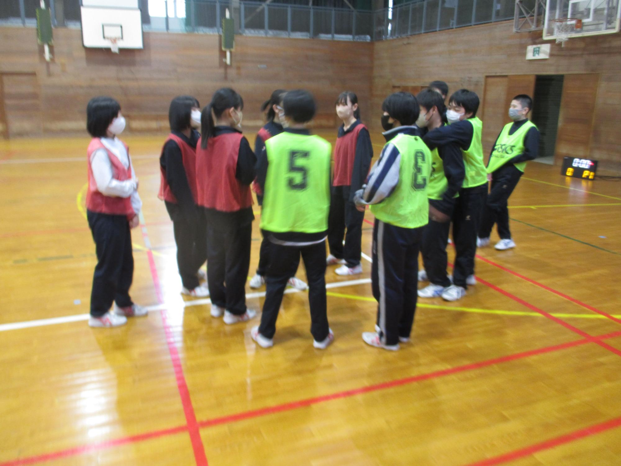 体育館で赤と緑のビブスを着た生徒たちが円になって話をしている写真